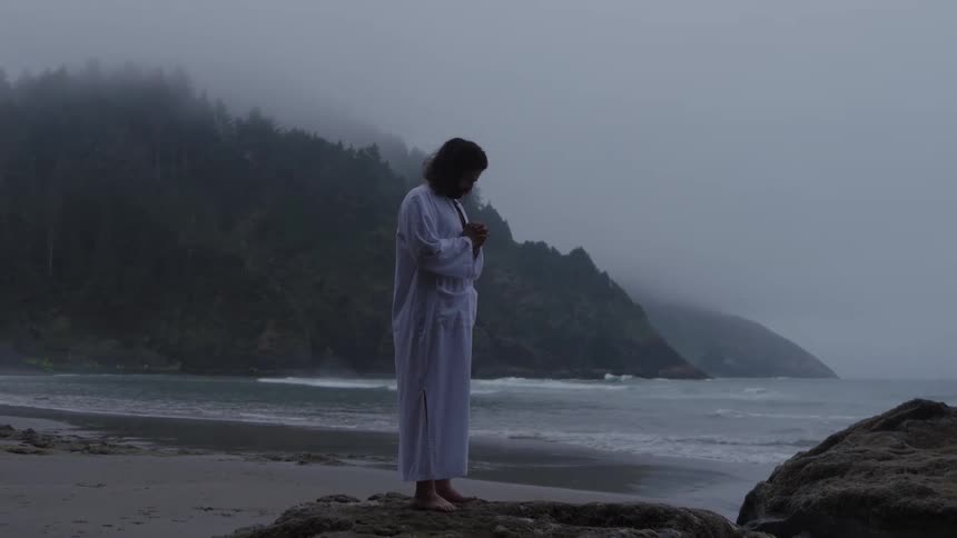 Man wearing a simple tunic stands on a coastal rock formation on a foggy day and prays