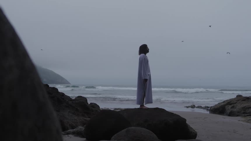 The video clip shows a distant shot of a person standing on a dark rock formation overlooking a foggy or overcast beach the person is silhouetted against the bright sky wearing a lightcolored long robe or dress and facing the ocean