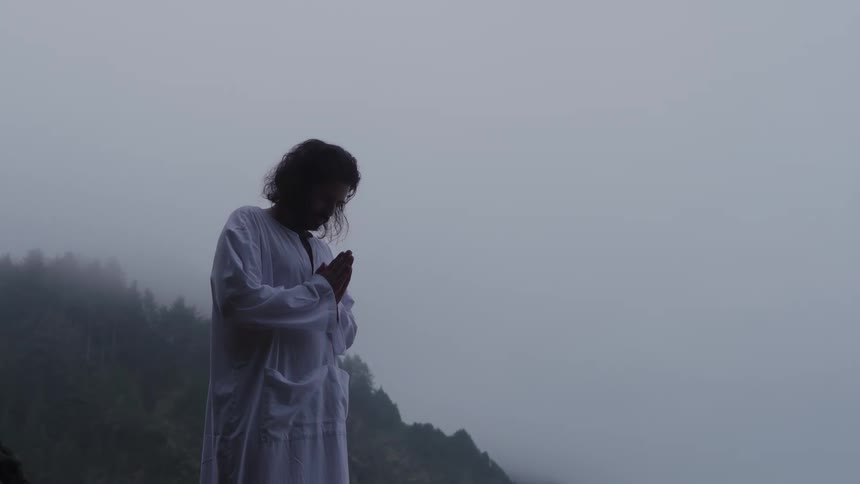 A man wearing a white longsleeved garment standing on a dark rocky shoreline with waves and foggy treecovered hills in the background