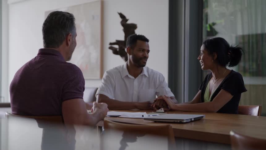 The video clip is a medium shot of a smiling diverse couple seated at a wooden table across from a man possibly a financial advisor or real estate agent the woman is holding the mans hand on the table suggesting a positive and collaborative dis