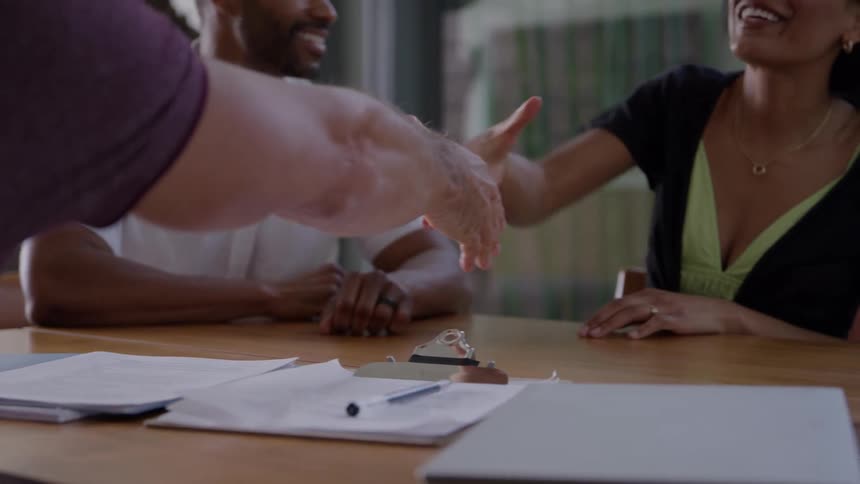Three people seated at a wooden table covered with paperwork where a man in a white shirt and an offcamera person in a purple sleeve shake hands across the table after which the seated man and woman both turn to each other and smile