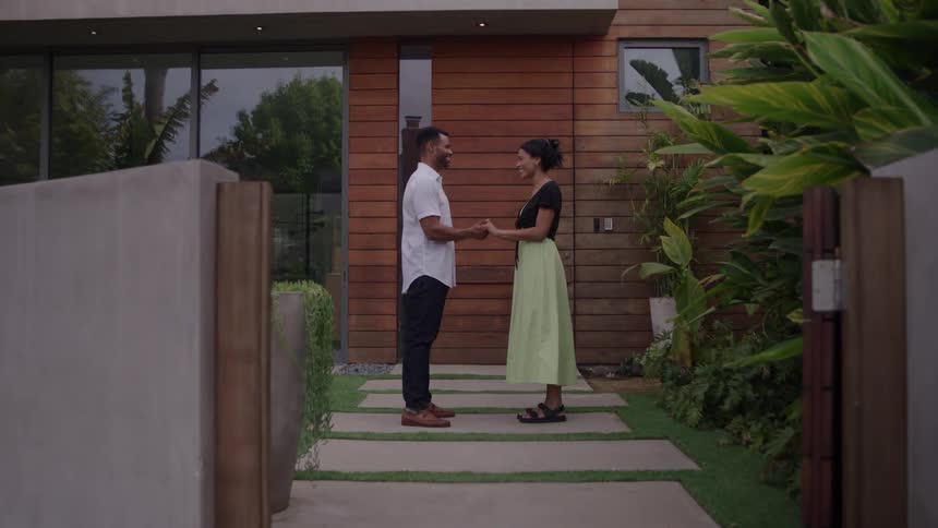 A happy couple embraces in front of their new modern home marked by a sold sign