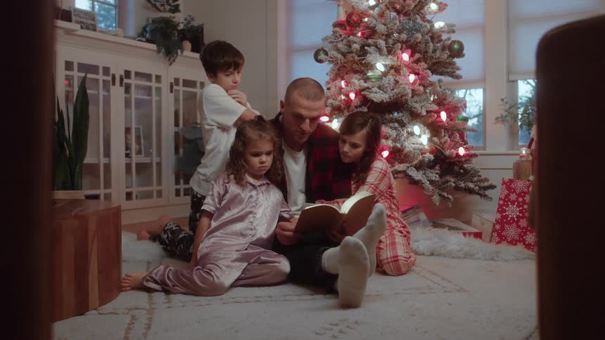 Man reading a book to three children sitting around a lit christmas tree