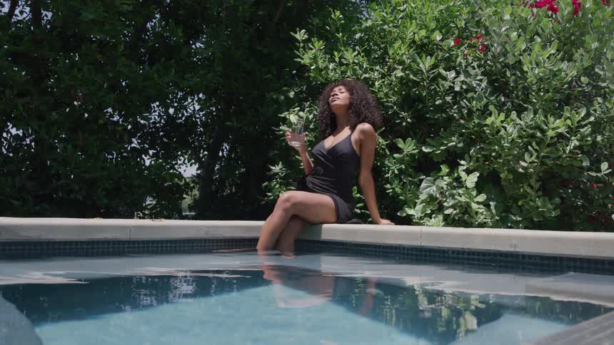 Woman holding a beverage sits at the side of a pool with her feet in the water on a summer day
