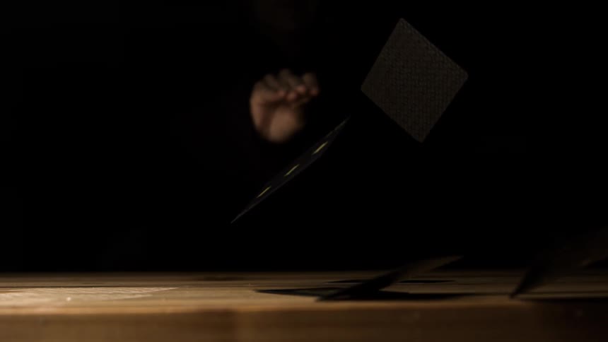 Person sitting in the dark drops black playing cards onto a wooden surface