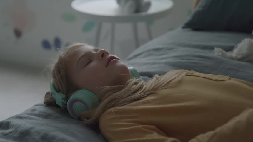 The video clip is a closeup of a young girl lying down with her eyes closed wearing a pair of light blue headphones she is wearing a yellow top and resting on a gray bedspread in a softly lit room