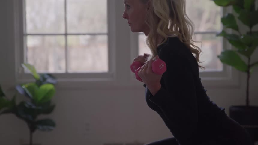 The video clip is a medium closeup of a persons torso and arms performing bicep curls with small pink dumbbells the person is wearing black athletic attire and standing indoors near a bright window