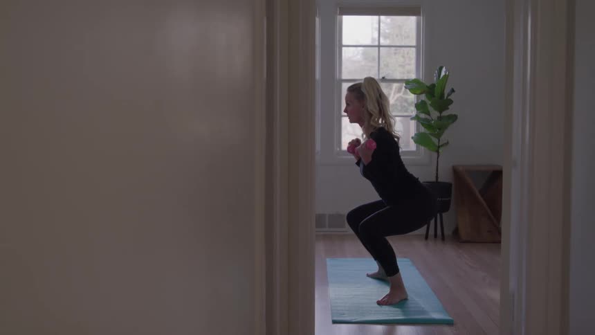 The video clip is a medium shot of a woman in black athletic wear performing a squat exercise while holding small pink dumbbells in a brightly lit room she is facing a window and standing on a blue exercise mat in what appears to be a home workout