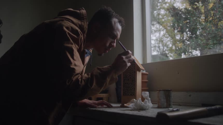 Man painting a small wooden house near a bright window