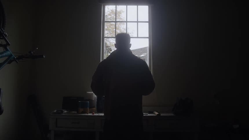 Man in a coat looking out the window in a dimly lit room