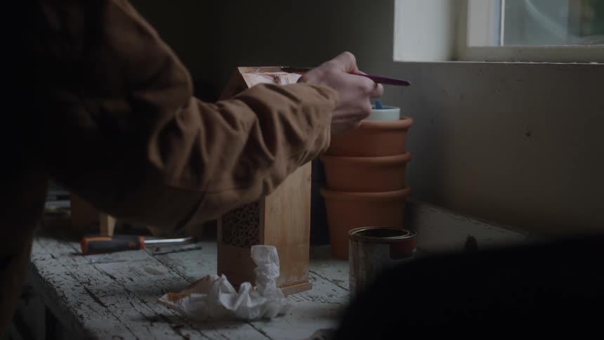 The video clip is a medium closeup of a persons hands carving or marking a small wooden box with a pencil the scene is indoors and dimly lit with craft materials and small terracotta bowls visible in the background