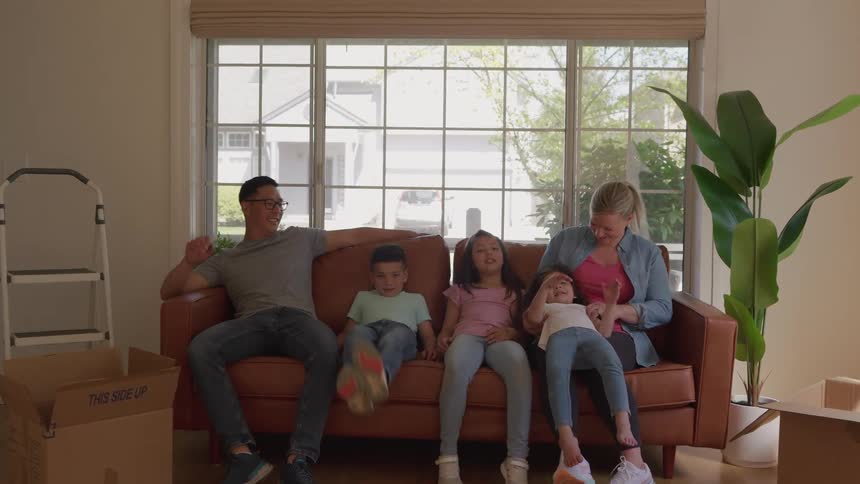 The video begins with a diverse family of five parents and three children standing formally in their new bright living room surrounded by moving boxesthe parents and kids then sit down together on the couch and relax and laugh symbo