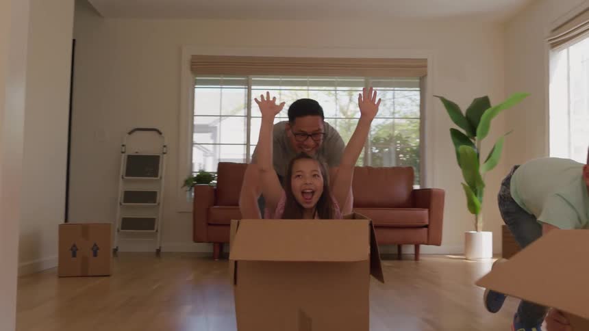 A joyful view shows a man and two girls laughing as they are pushed in large cardboard moving boxes by another boy in a brightly lit unfurnished living room with hardwood floors