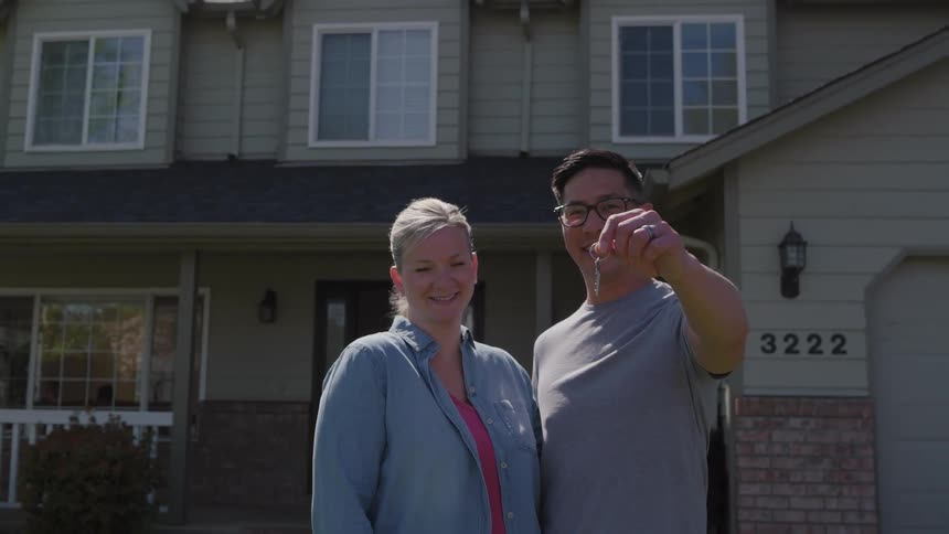 The video features a medium shot of a smiling diverse couple standing proudly in front of a twostory suburban housethe man holds up a set of keys toward the camera symbolizing a new home purchase or real estate achievement