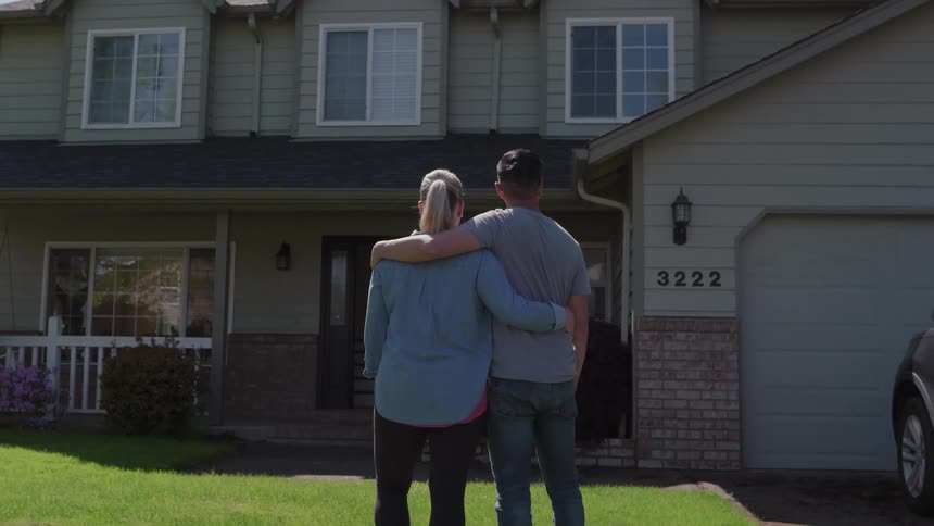 The video is a wide shot of a diverse couple seen from the back standing on a green lawn in front of a twostory suburban housethey are sidebyside with arms around each other looking at the house symbolizing pride and ownership