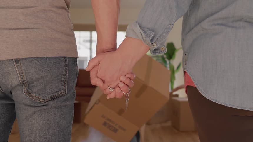 A man and woman standing sidebyside holding clasped hands between them with a set of silver keys dangling from their interlocked fingers as they stand in a room with some kids playing with cardboard boxes