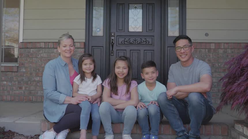 The video is a mediumwide slightly lowangle shot of an asiancaucasian couple and their three children sitting on the steps outside their homethe family is smiling directly at the camera conveying happiness and togetherness in front of thei