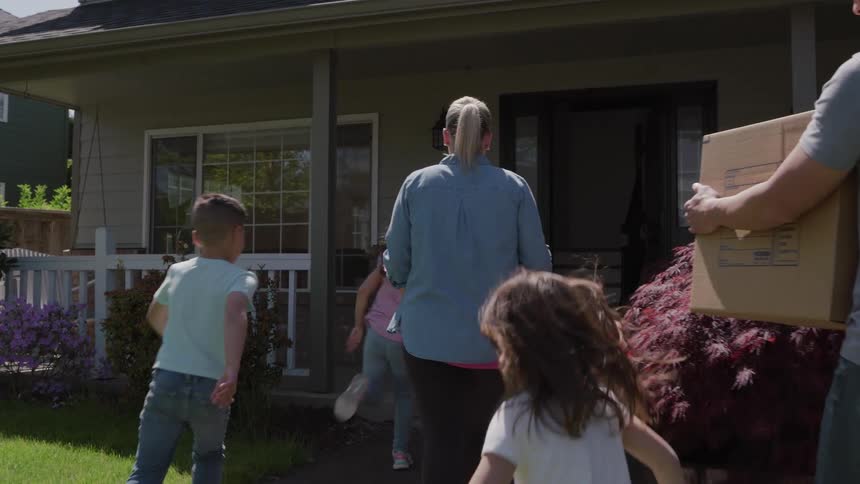 An adult man and woman walking towards a door of a house and three children are running in front of them
