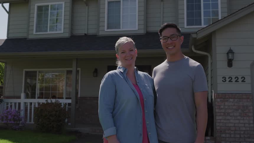A man and a woman are standing smiling as they look forward and behind there is a large house
