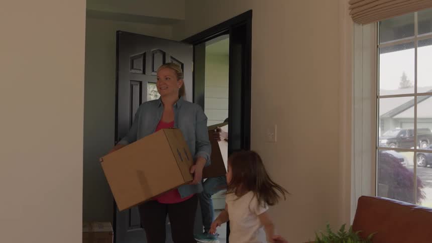 The video shows two children a girl with a large cardboard box and a boy behind her walking through the open front doorway of a housethey both smile happily as they carry their boxes into the bright interior suggesting they are moving in