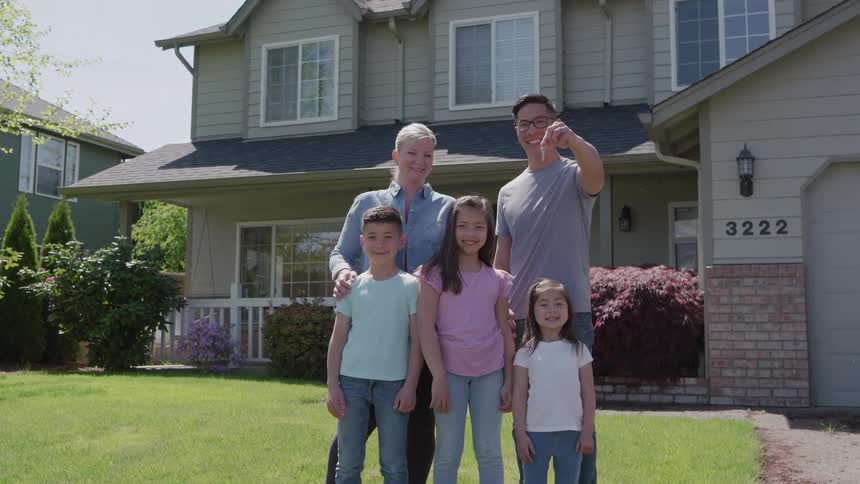 A smiling mixedrace family of five including a man and a woman with three children stands outside their light green suburban house on a sunny day holding up a new house key