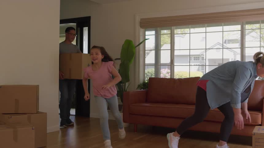 Five people entering a room through a doorway with the mother and father carrying cardboard boxes and the children running past them as they move their belongings into a new bright and mostly empty living room