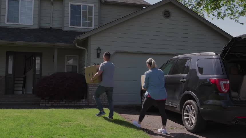 A couple carries moving boxes from the back of an suv towards the front door of their suburban home