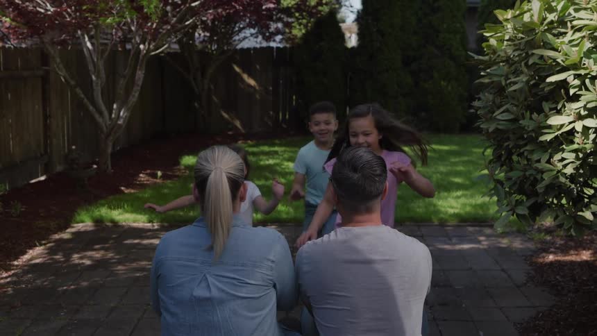 Three children running across a grassy lawn toward two adults seated on a paver patio culminating in the children hugging and surrounding the parents in a central family embrace in a suburban backyard