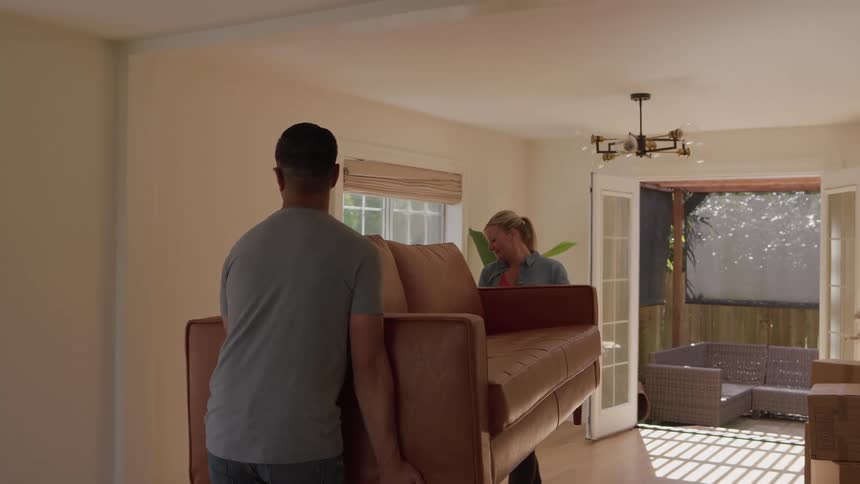 The video shows a couple carefully carrying a large brown leather couch into a bright empty room with large windowsthey slowly navigate the living space and eventually move out of view into an interior room likely moving into a new home or red