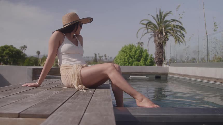 A woman wearing a straw hat and a lightcolored outfit sitting on a wooden deck with her legs extended and her feet dipped into the water of a small modern swimming pool on an outdoor terrace with tropical plants