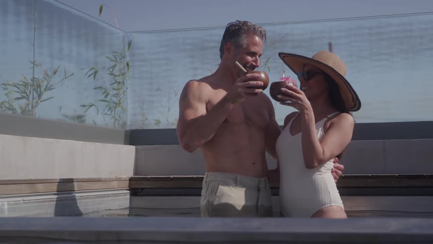 A middleaged man and a young woman in swimsuits and sun hats hug and cheer with coconut drinks standing in an outdoor pool on a sunny day
