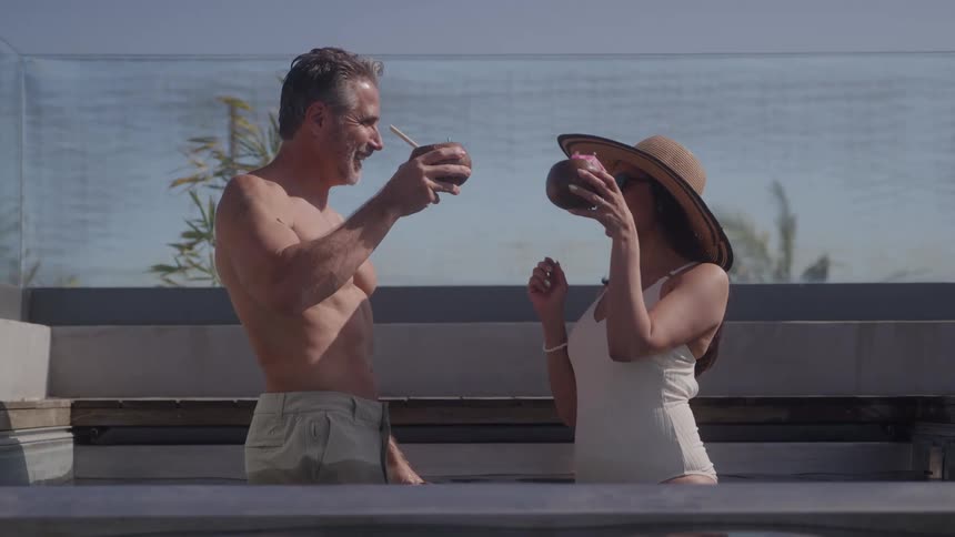The video is a medium shot of a middleaged shirtless man and a woman in a white swimsuit and sunhat standing in an outdoor hot tubthey are smiling and clinking tropical drinks served in coconut shells suggesting a moment of vacatio