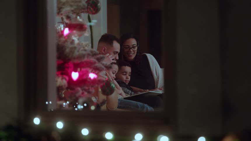 Family gathered on the couch reading a book near the glowing christmas tree