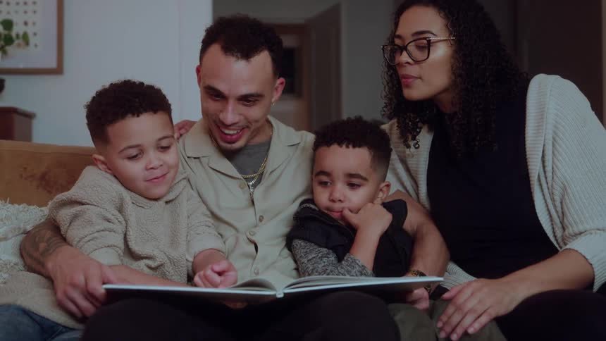 Two adults and two young boys sitting closely together on a couch with the father holding an open book in front of them as they look down at the pages and smile in a dimly lit living room