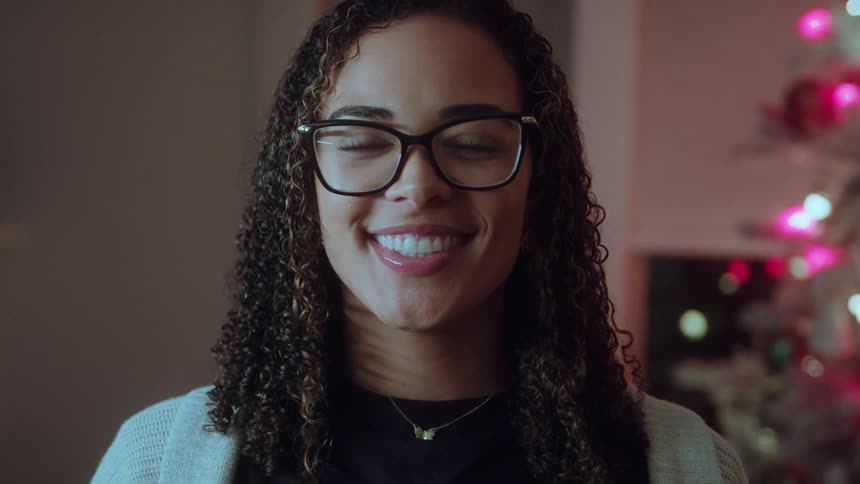 A young woman with long curly hair and eyeglasses who is smiling broadly directly into the camera with outoffocus holiday lights visible in the dark background