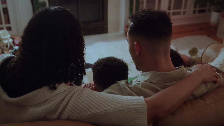The video is a rearview closeup shot of a couple sitting on a sofa with their small child nestled between themthey are in a dimly lit cozy living room possibly during the holidays given the faint festive decorations and are looking out
