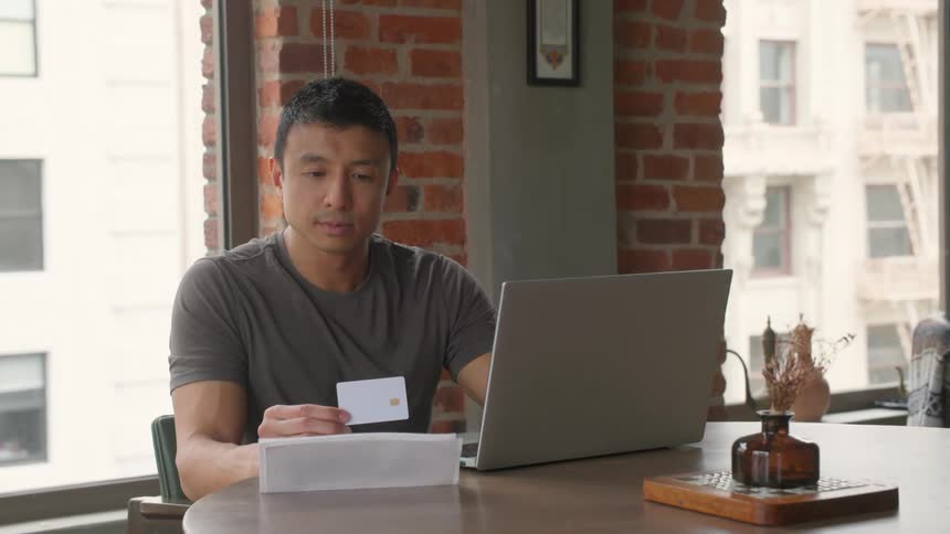 Man sitting at a table holding a card while using a laptop near the window