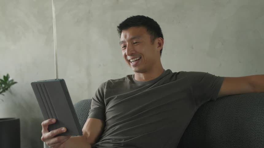 The man is sitting on the couch and laughing at his tablet