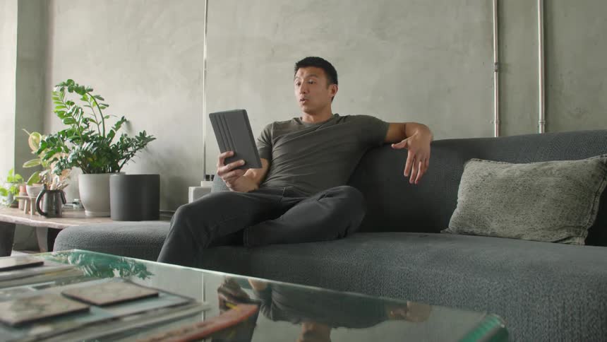 Man sitting on his couch communicates through a video call on his tablet during the daytime