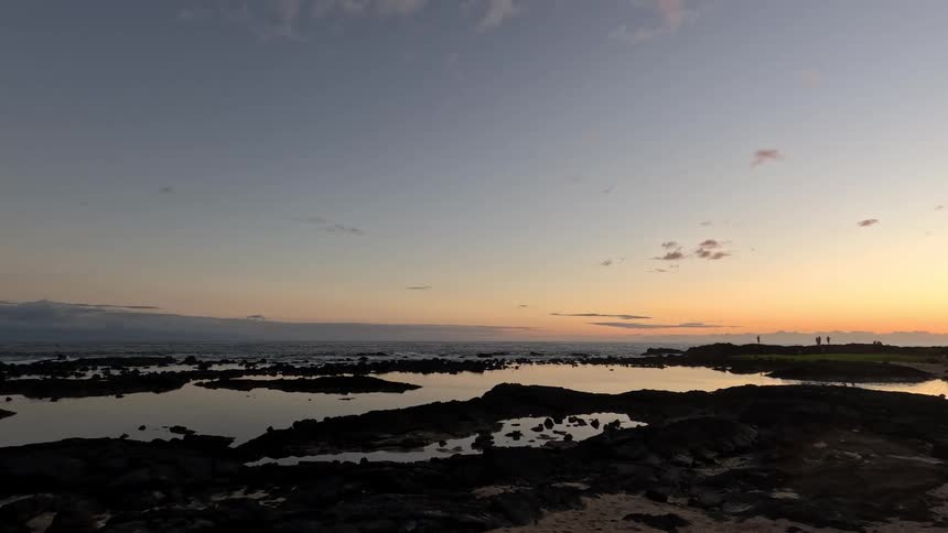 The video is an establishing shot of a dark rocky shore against a colorful sunset or sunrise skythe still pools of water reflecting the light and the dark landscape suggest a dramatic tranquil setting at the transition of day and night