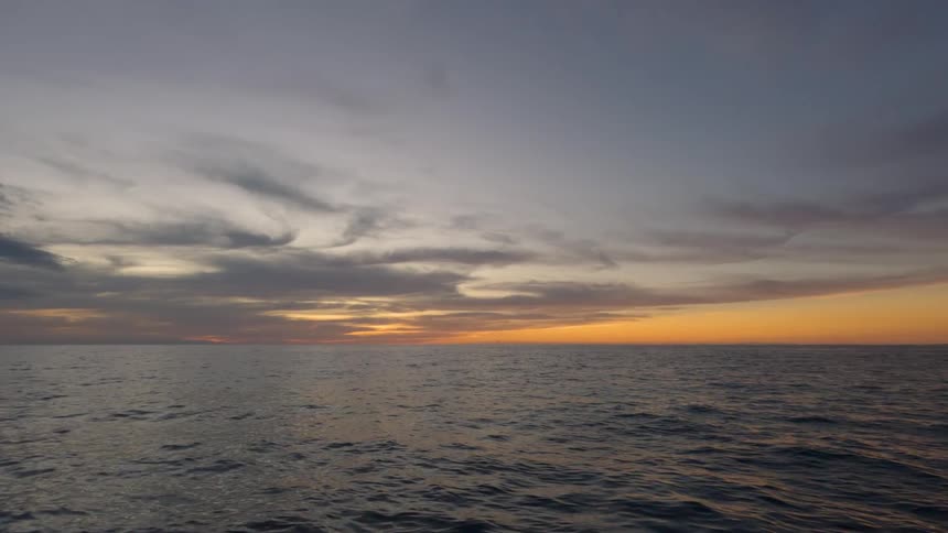 A wide view captures the dark wavy ocean water under a dramatic sky with gray clouds and a bright orange glow along the horizon at sunset or sunrise