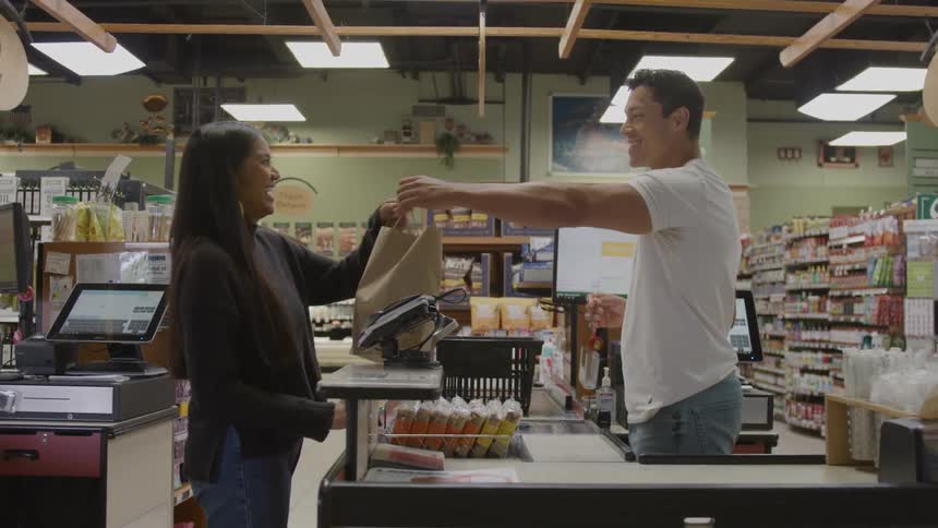 The video shows a medium shot of a man and a woman interacting at a supermarket checkout counterthe man acting as the checker hands a paper bag of groceries to the smiling woman suggesting a pleasant transaction