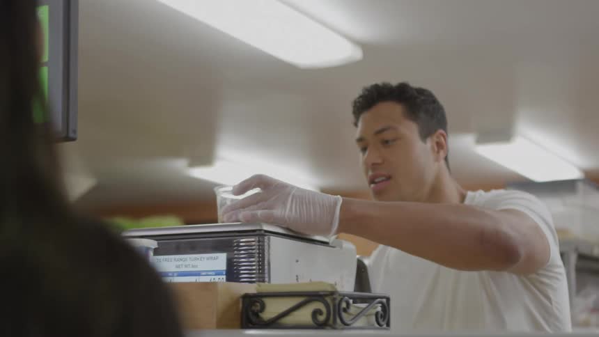 A food service worker wearing white gloves who scoops an ingredient from a bowl into a plastic container behind a glass counter then places the container on a digital scale before looking up and speaking to a customer across the counter