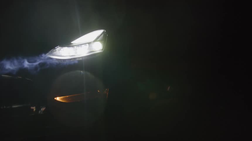 A bright led car headlight cuts through the darkness illuminating streaks of smoke