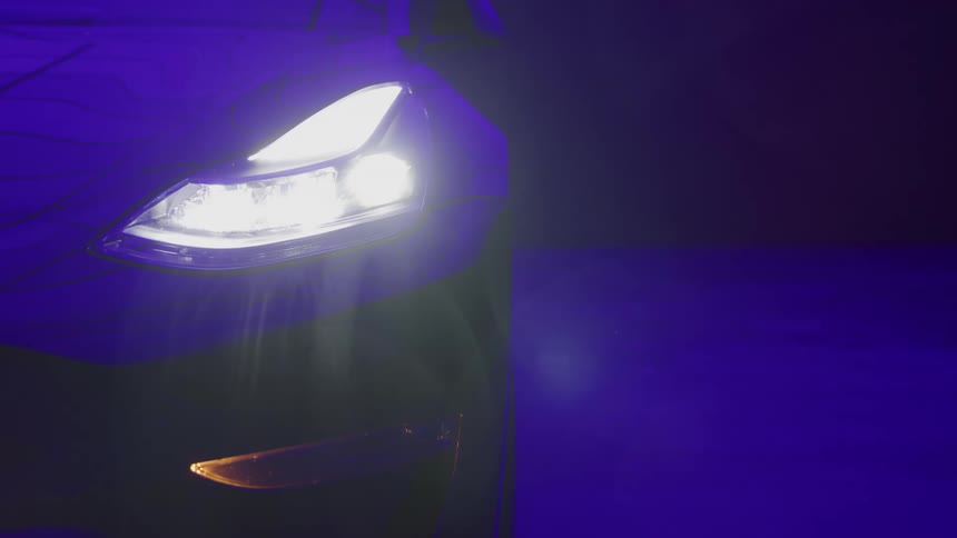A bright white headlight and corner of a modern car in a dark environment with the intense beam casting a wide illumination on the immediate area and the adjacent vertical surface reflecting a dark blue light