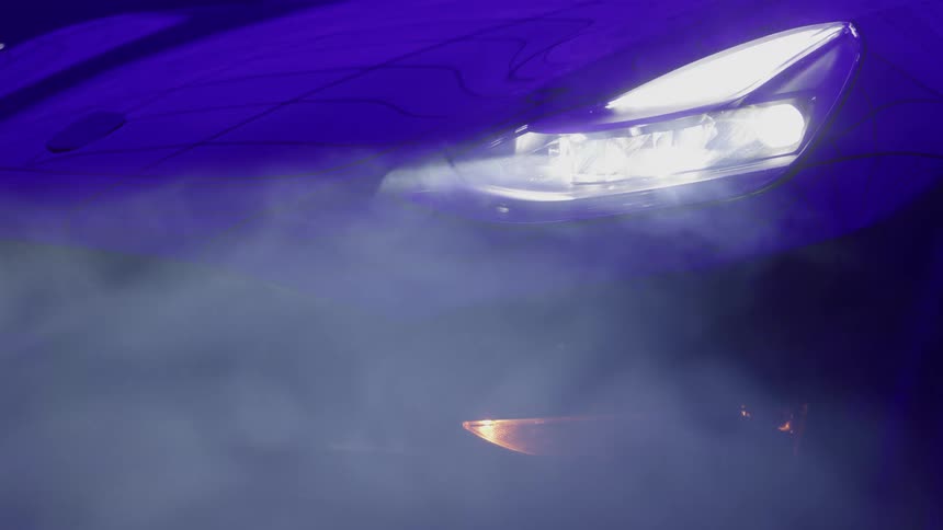 The video is a stylized closeup shot of the front of a sports car or luxury vehicle at nightits bright headlight cuts through the surrounding blue light and wisps of fog or smoke creating a dramatic highcontrast effect