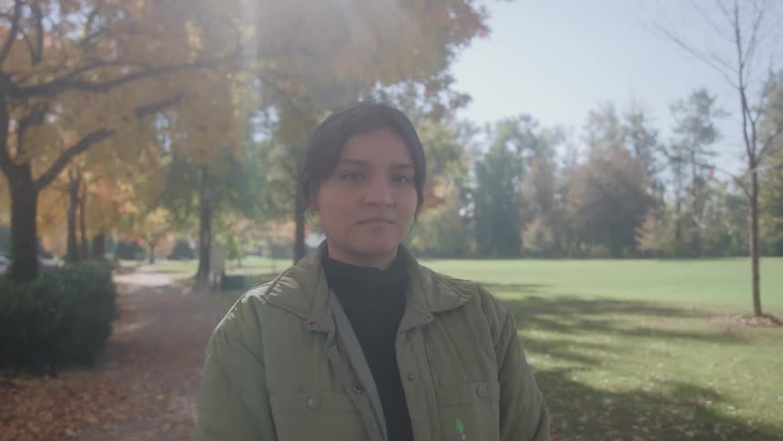 A young woman with dark hair wearing a green jacket and black top looking directly at the camera as she stands on a treelined park path in the midst of bright autumn foliage
