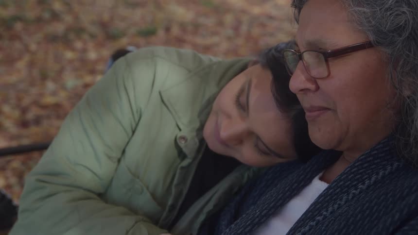 Woman resting her head on her mothers shoulder while sitting on a park bench