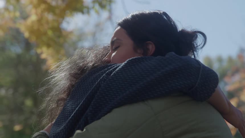 The video is a closeup shot of a darkhaired woman being embraced in a deep tender hug by another person outdoors on a sunny dayshe has her eyes closed and her face pressed into the other persons shoulder conveying a sense of strong emoti