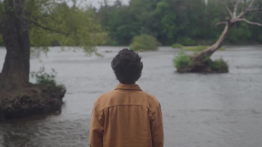 A wide view from behind shows a person with dark curly hair wearing a brown jacket standing at the edge of a calm river on an overcast day with lush green forest in the distance